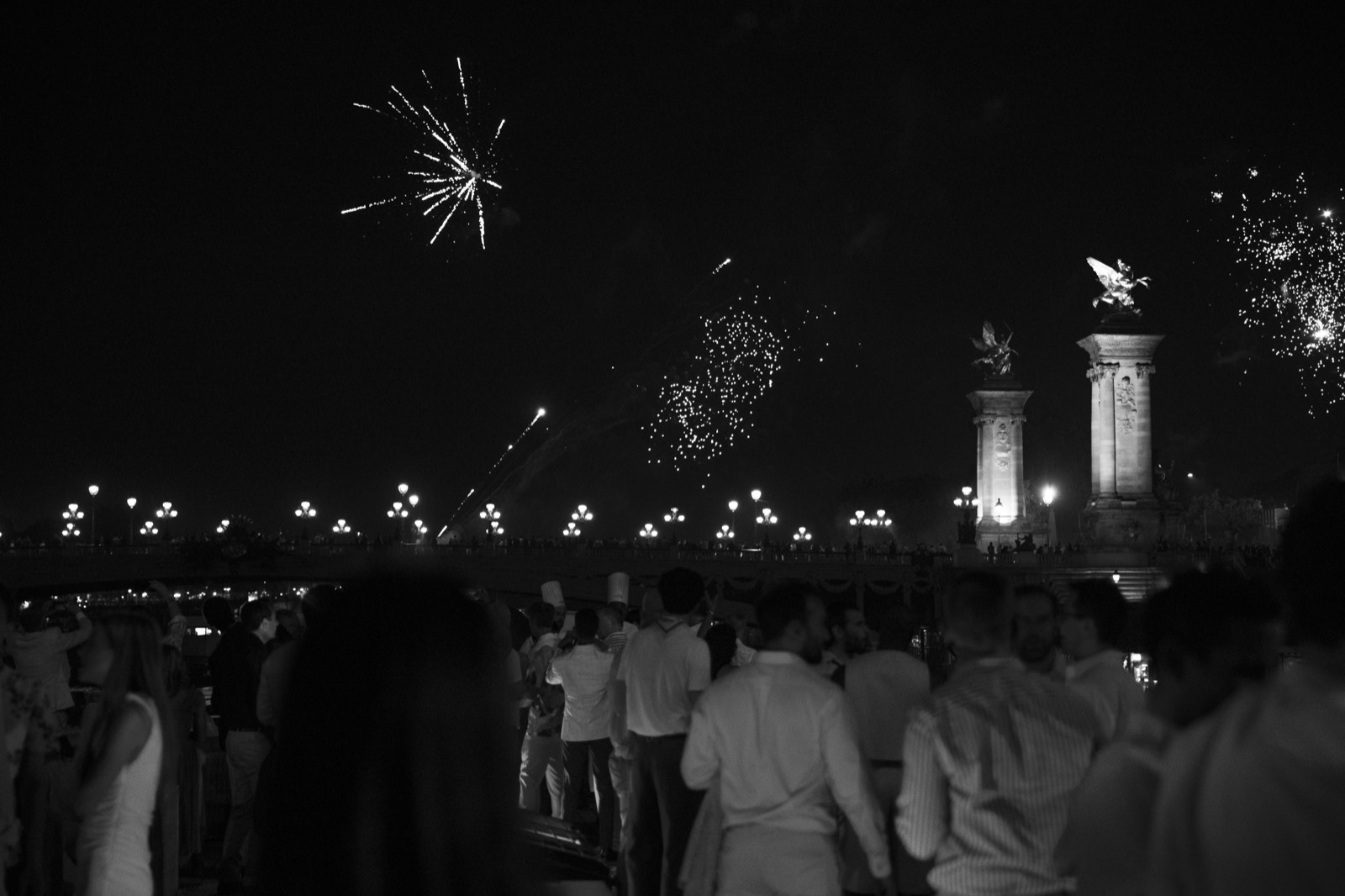 Black and white view of fireworks over Pont Alexandre III with guests in white attire watching