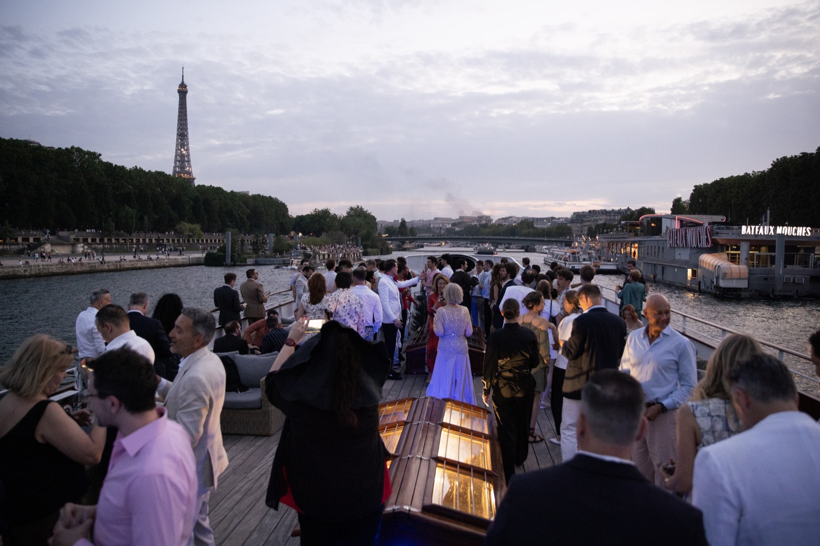 Guests in elegant evening attire on the yacht's deck, the Eiffel Tower glowing in the distance at dusk