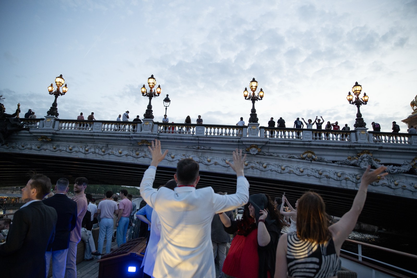 Guests celebrating with raised arms as the yacht passes beneath the illuminated Pont Alexandre III